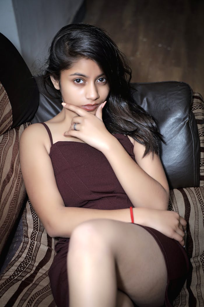 Home Young woman sitting and posing on a couch in a stylish brown dress indoors.